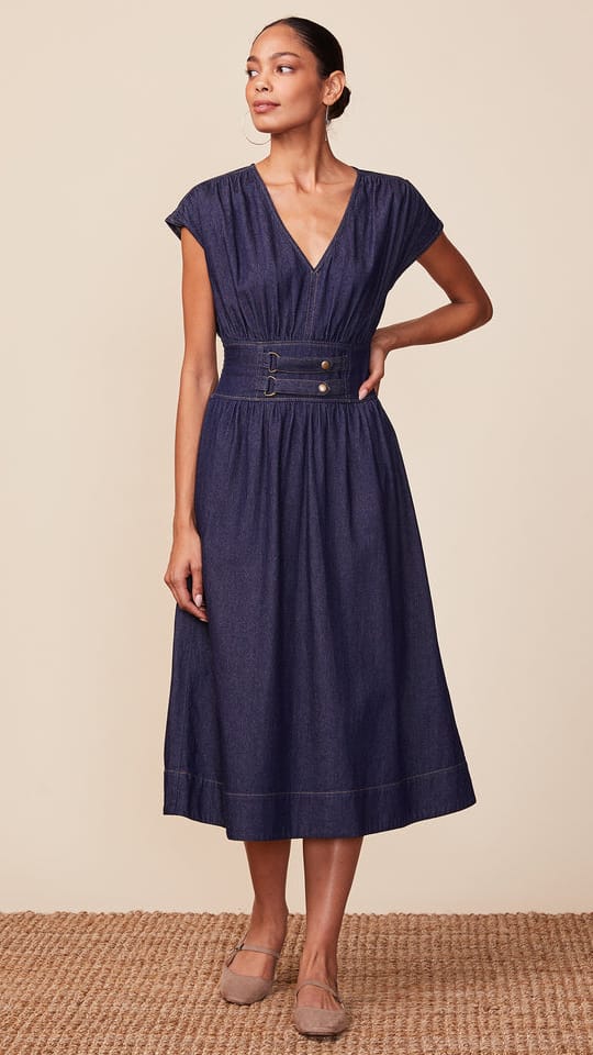 A woman models a navy blue midi dress with cap sleeves, a V-neckline, a matching fabric belt with gold-tone buttons, and a pleated skirt, set against a neutral beige background.