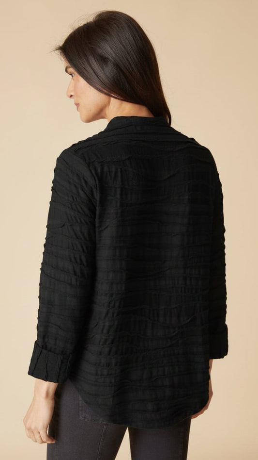 A woman shown from the back wearing a black textured, vertically pleated long-sleeve blouse against a solid beige background.
