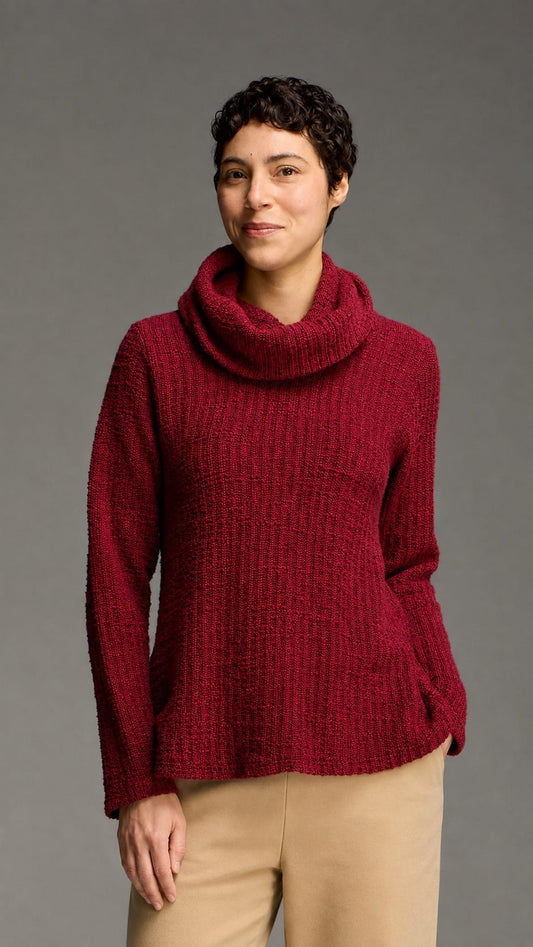 A woman wearing a chunky knit cowl-neck sweater in deep burgundy, paired with beige pants, standing against a solid gray studio background.