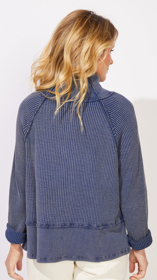 A woman shown from the back wearing a textured, blue-and-white vertically patterned long-sleeve top with rolled cuffs, paired with cream pants, against a plain white studio background.