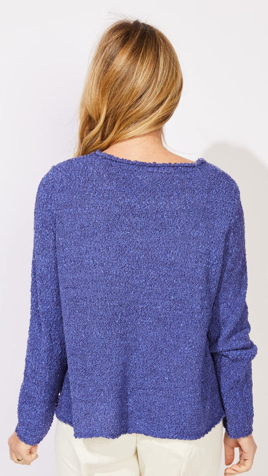A woman viewed from behind wearing a textured, heathered blue-purple knit sweater and cream pants against a plain white background.