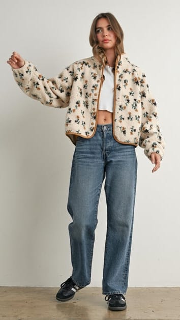 A woman modeling a cropped, cream-colored sherpa jacket with a black floral pattern and tan trim, paired with blue jeans and black shoes, against a plain light gray wall.