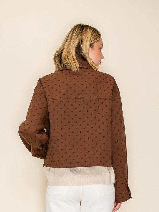 A woman shown from the back wearing a brown cropped jacket with a small repeating polka dot pattern, layered over a cream knit top and white pants, against a plain off-white background.