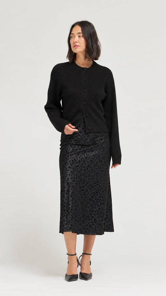 A woman modeling a black long-sleeve button-front cardigan and a black textured midi skirt with black pointed-toe heels, photographed against a plain light gray studio background.