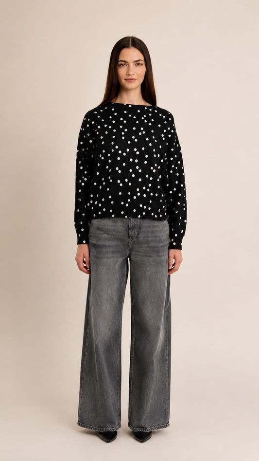 A woman stands against a plain beige studio background wearing a black long-sleeve top with white polka dots and wide-leg gray jeans.