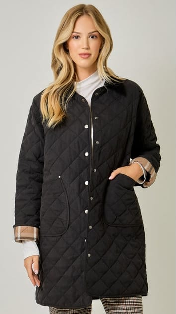 A blonde woman models a black diamond-quilted button-up coat over a white turtleneck, showing plaid lining on the cuffs, against a light gray studio background.