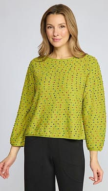 A woman modeling a lime green long-sleeve knit top with a small multicolor speckled pattern, paired with black pants, against a light gray studio background.