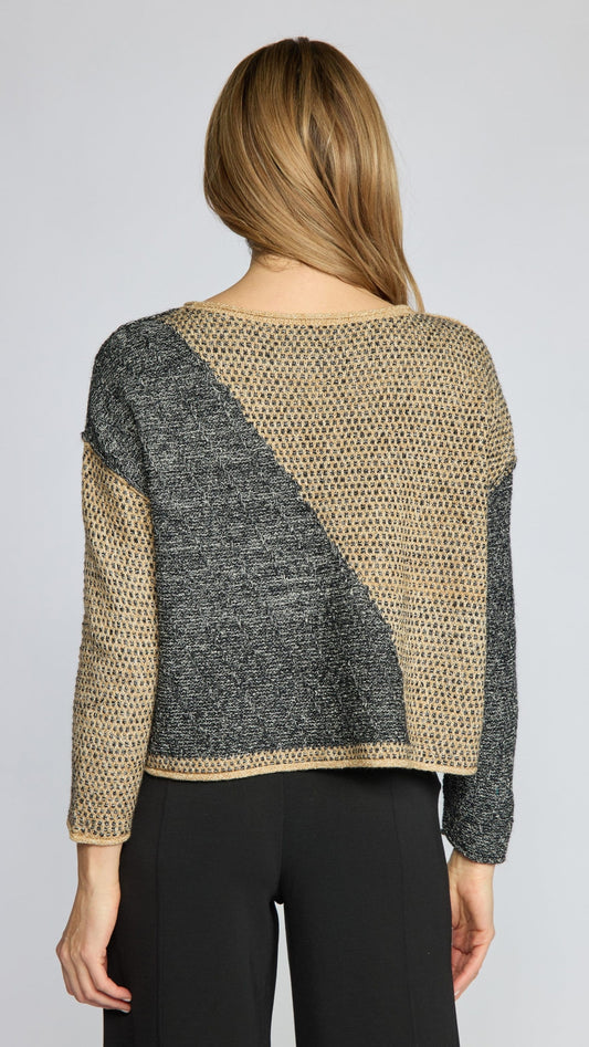 A woman viewed from the back wearing a cropped knit sweater with a diagonal color-block pattern in beige and dark gray, paired with black pants, against a light gray studio background.