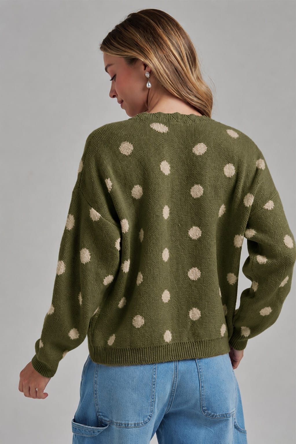 A woman shown from the back wearing an olive green knit sweater with beige polka dots, paired with light blue jeans, against a solid gray studio background.