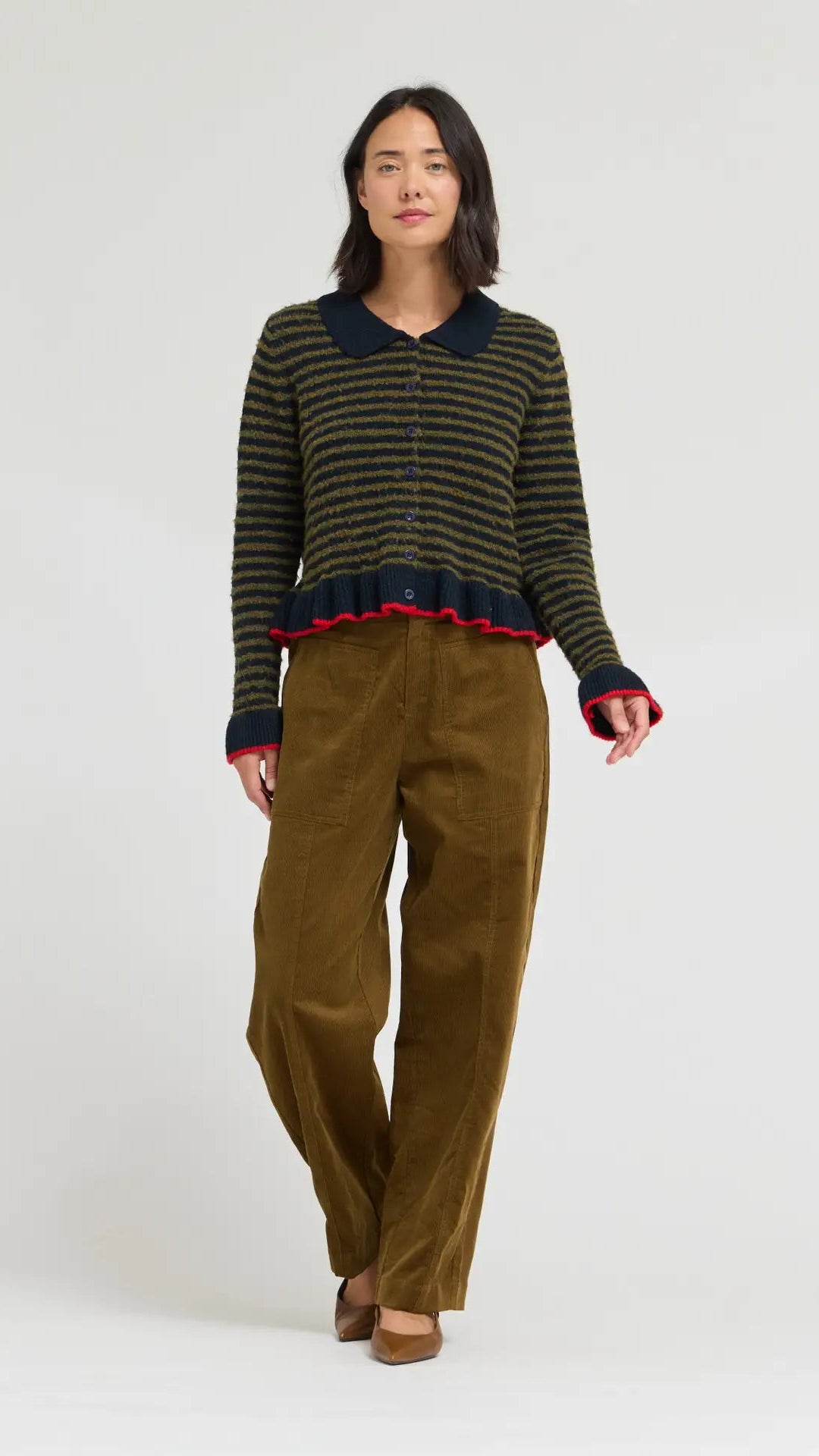 A model stands against a light gray studio background wearing a dark striped knit cardigan with a navy collar and red trim, paired with olive-brown wide-leg trousers.