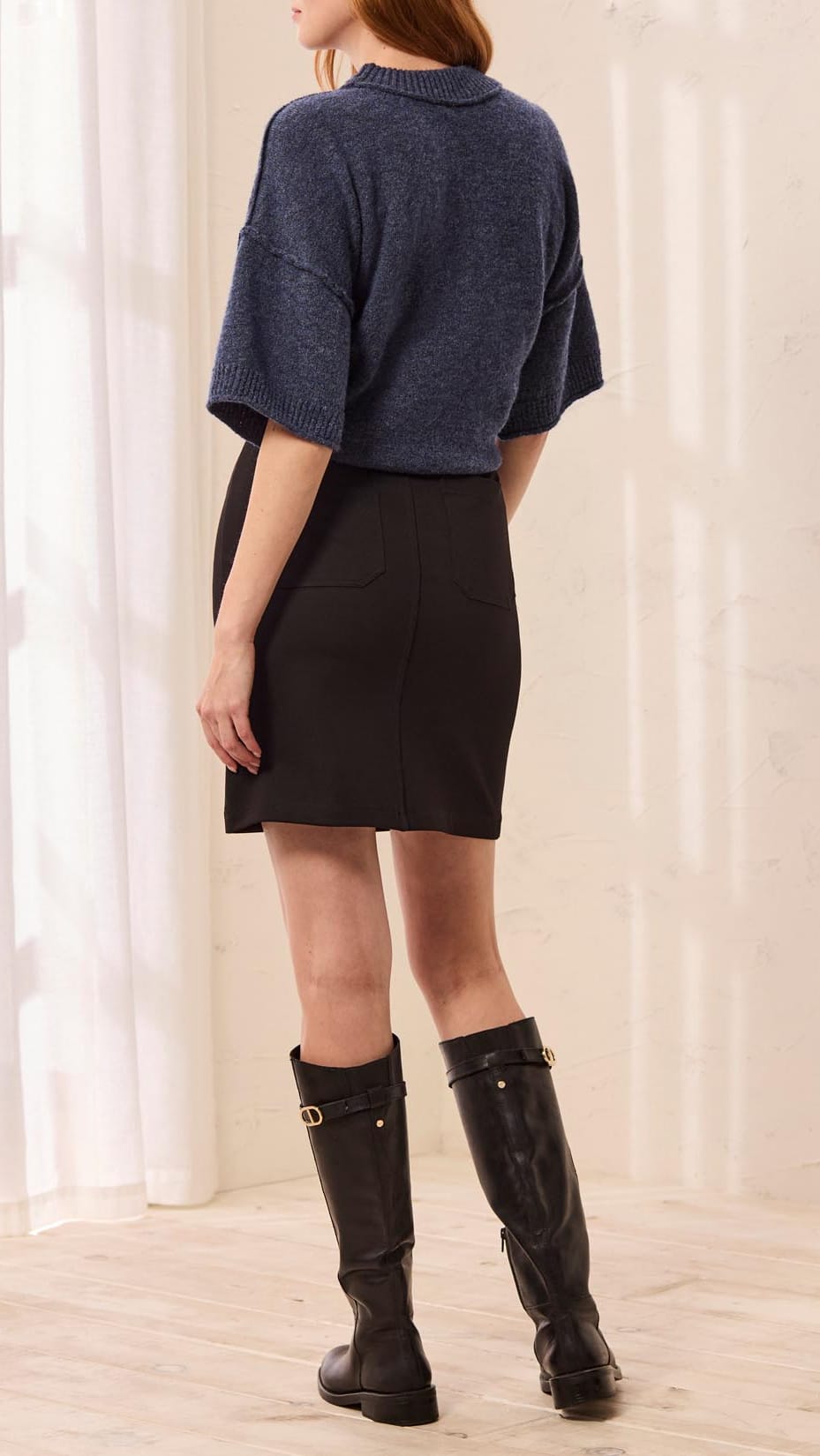 A woman is shown from behind wearing a dark blue knit short-sleeve top, a black mini skirt, and black knee-high boots in a brightly lit room with white curtains.