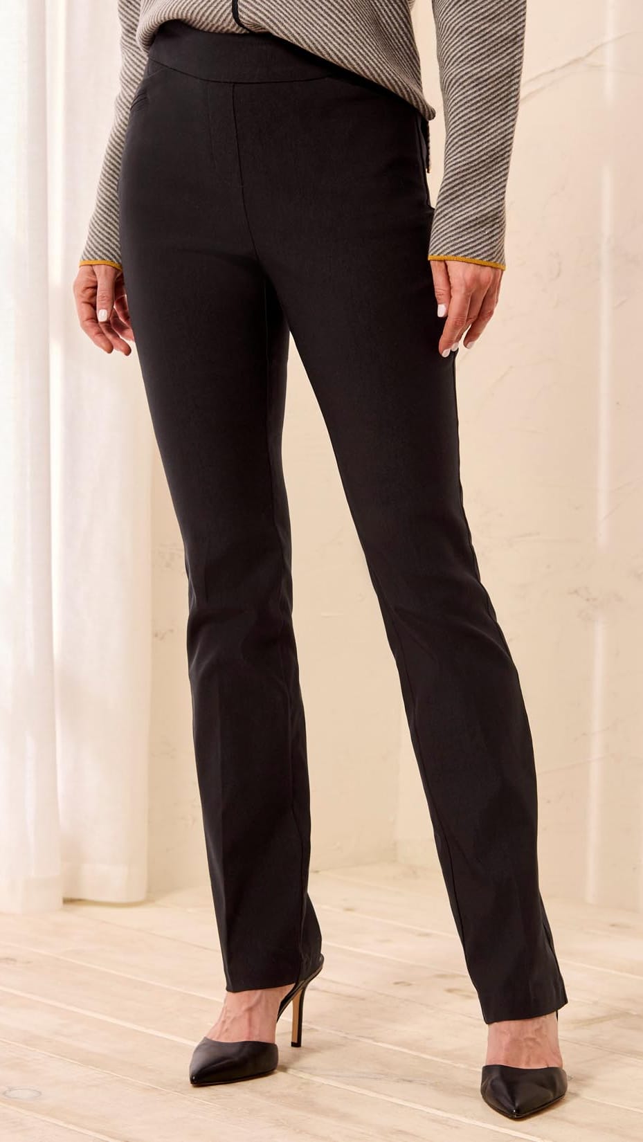 A person wearing black straight-leg trousers, a gray and white striped long-sleeve top, and black pointed-toe heels, standing in a brightly lit indoor setting with light wood floors and white curtains.