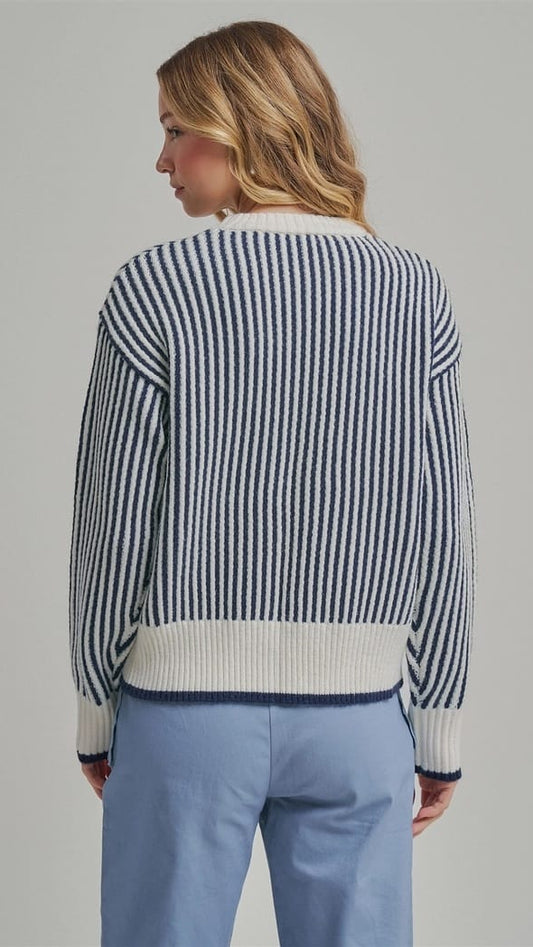 A woman shown from the back wearing a navy and white vertically striped knit sweater with solid white ribbed trim, paired with light blue trousers, against a solid gray studio background.