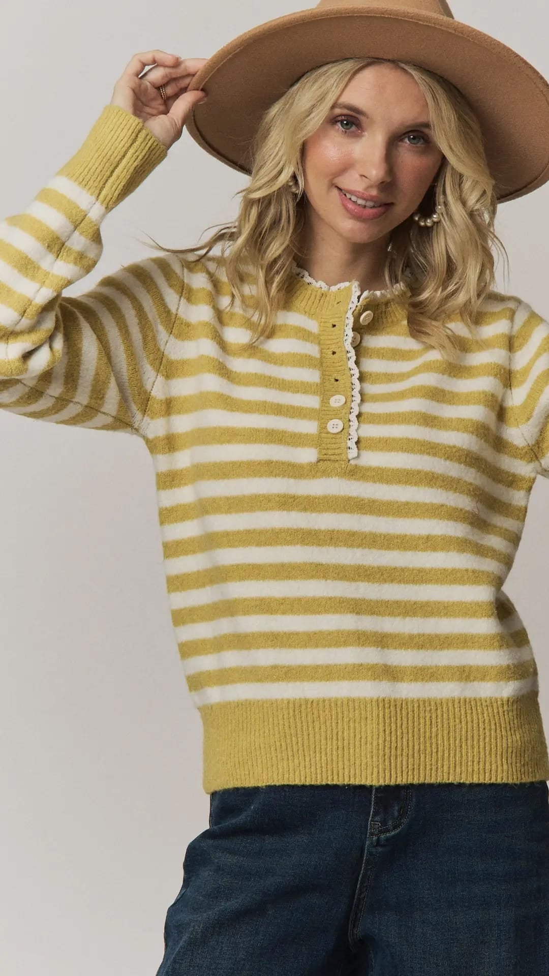 A woman wearing a yellow and white striped knit henley sweater, a wide-brim tan hat, and dark blue jeans, posing against a light gray studio background.