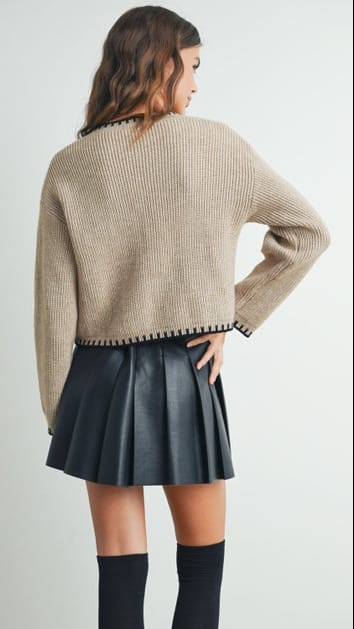 A woman shown from the back wearing a beige ribbed knit cropped sweater with black trim, a black pleated mini skirt, and black knee-high socks against a light gray studio background.