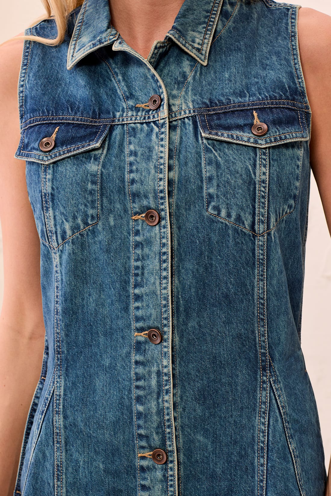 Close-up of a sleeveless blue denim shirt dress with a pointed collar, button front, and flap chest pockets, shown on a model against a light neutral background.