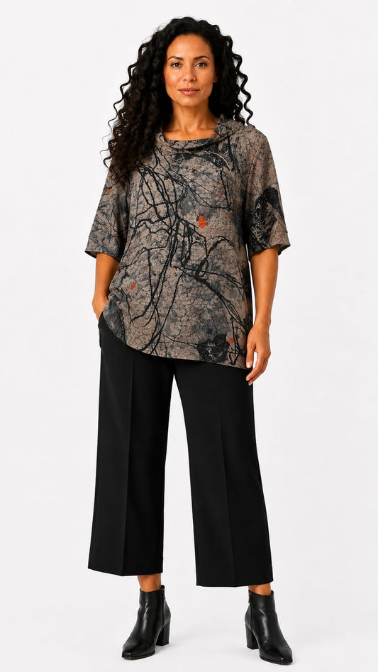 A woman modeling a short-sleeve top with a dark abstract branch-like print in gray, black, and muted orange, paired with black cropped pants and black ankle boots, against a plain light gray studio background.