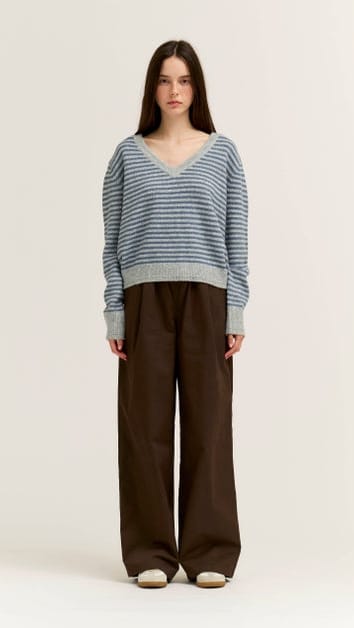 A model stands against a plain light background wearing a blue and gray striped knit sweater with wide-leg brown pants and white shoes.
