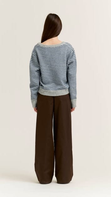 A woman shown from the back wearing a blue and gray striped long-sleeve sweater and dark brown wide-leg pants, standing against a plain off-white studio background.