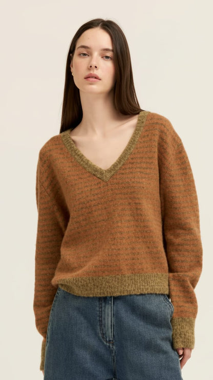 A woman wearing a brown ribbed knit sweater with olive green trim on a V-neck and hem, paired with blue jeans, against a plain light background.