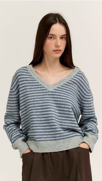 A woman wearing a blue and gray striped knit sweater with a wide V-neck, paired with dark brown pants, against a plain light background.