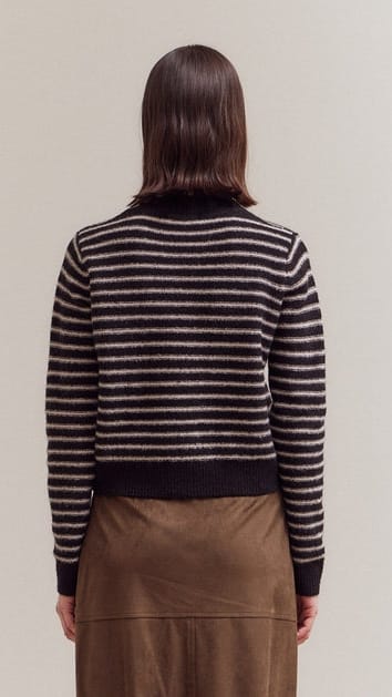 A woman viewed from the back wearing a black and white horizontally striped knit sweater and a brown skirt, against a plain light beige studio background.