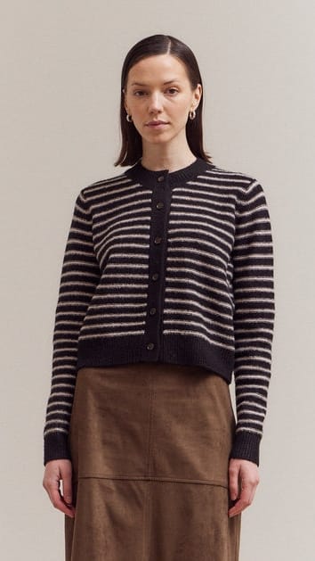 A woman modeling a cropped, long-sleeve knit cardigan with horizontal black and beige stripes, featuring a dark button-up front, paired with a brown skirt against a plain light gray studio background.