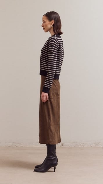 A woman stands in profile wearing a black and white horizontally striped knit sweater, a brown midi skirt, and black pointed-toe heeled boots against a plain light beige studio wall and floor.