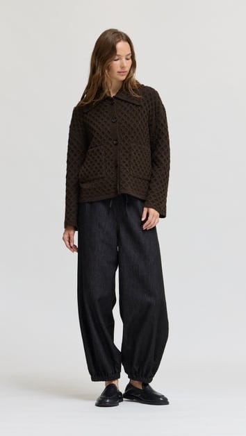 A woman modeling a dark brown textured cropped jacket with black wide-leg trousers and black loafers, standing against a light gray studio background.