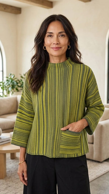A woman wearing a green and yellow striped mock-neck top with three-quarter sleeves and a front pocket, standing in a modern living room setting.