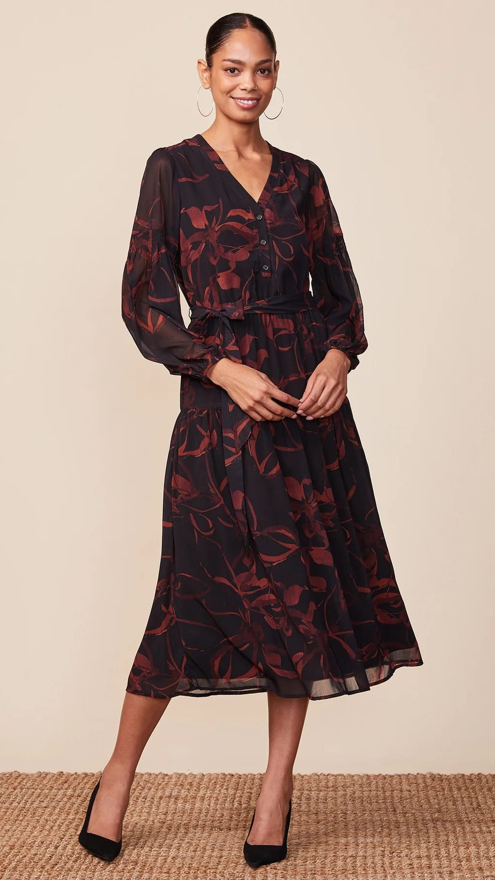 A woman modeling a long-sleeve midi dress with a dark base and a reddish-brown floral pattern, paired with black heels against a neutral beige background.