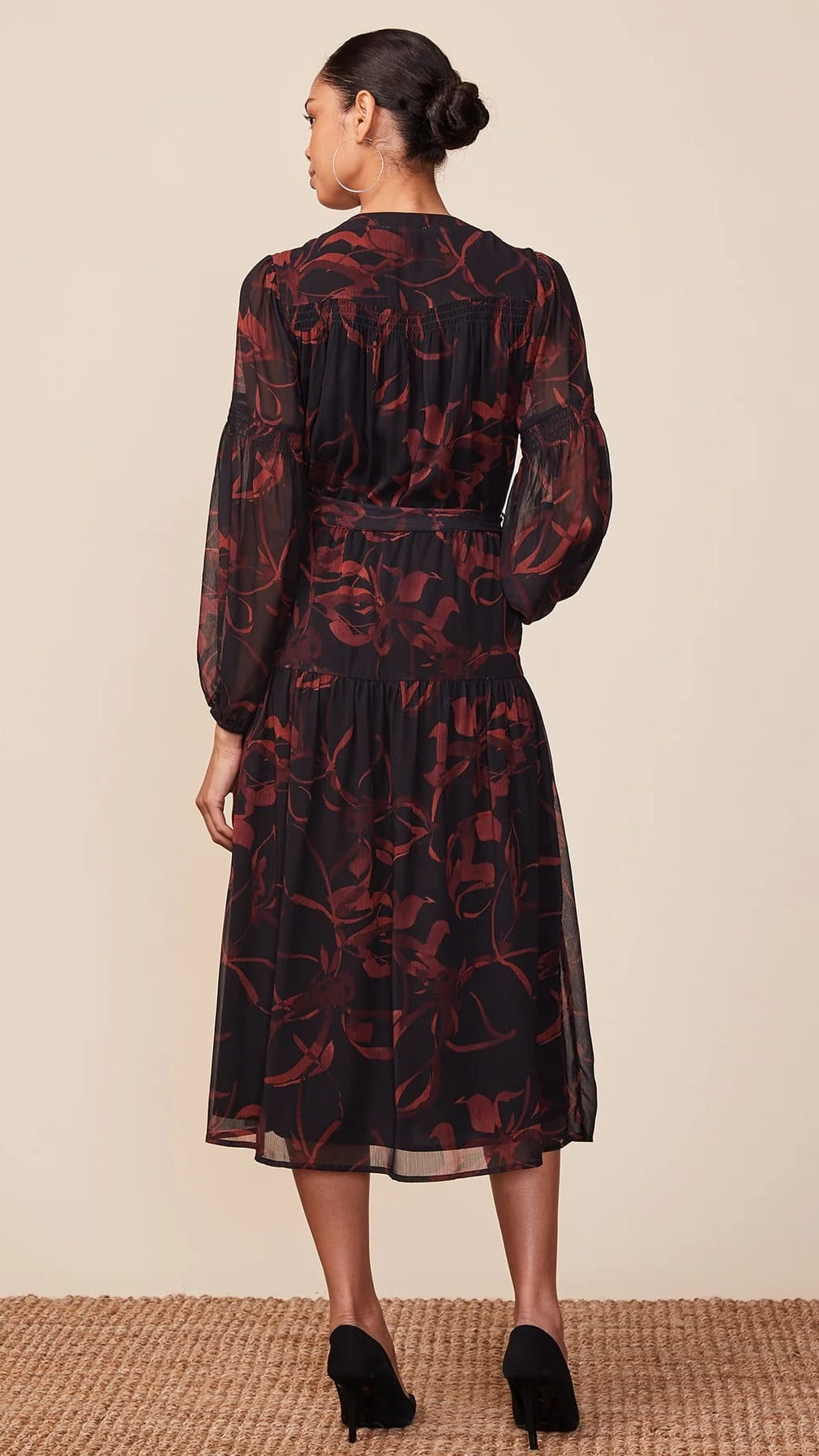 A woman viewed from the back wearing a long-sleeve midi dress with a sheer black base and a dark red floral pattern, paired with black high heels against a neutral beige background.