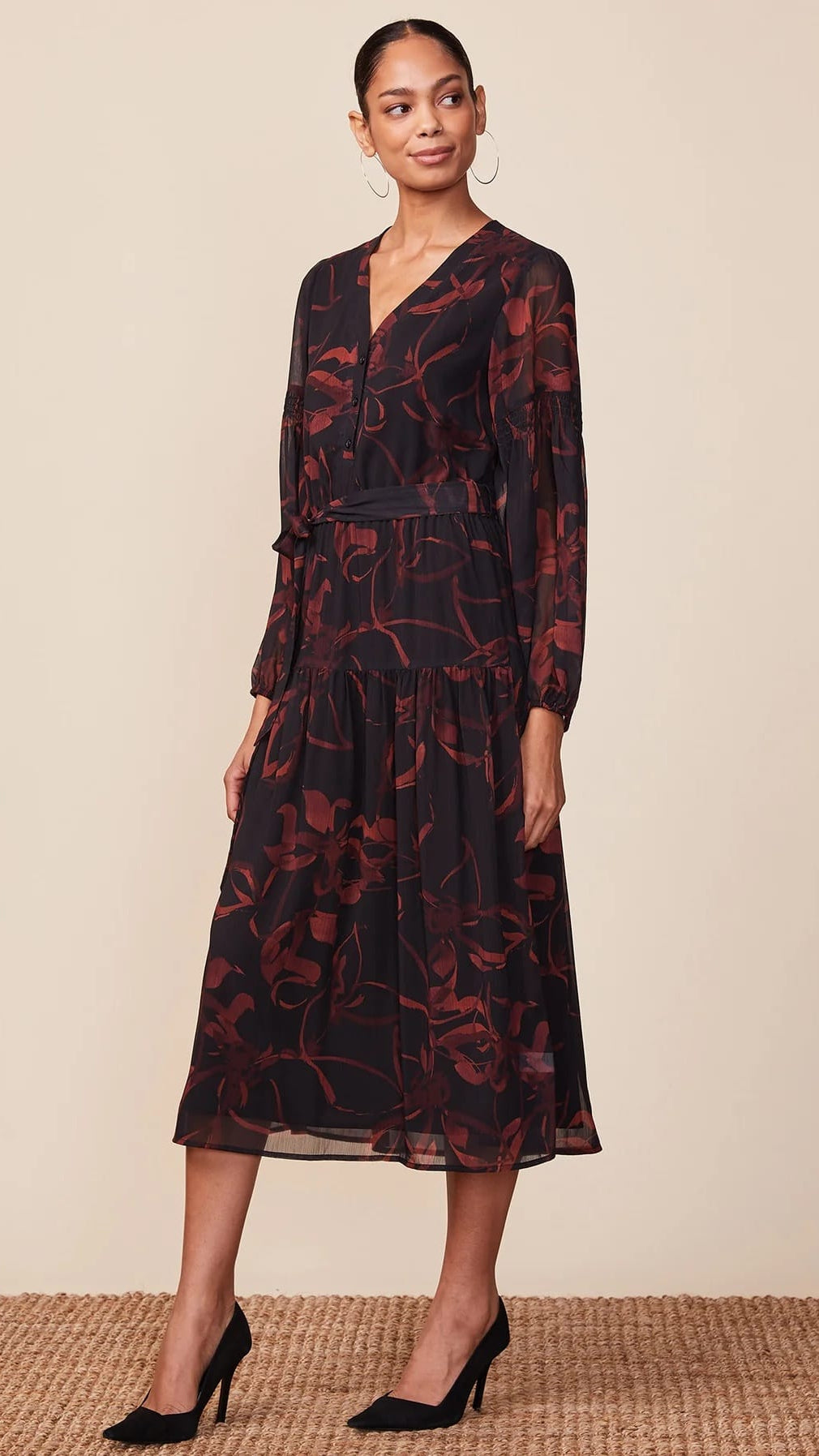 A woman modeling a long-sleeve midi dress with a dark base and red floral pattern, paired with black pointed-toe heels, against a neutral beige background.