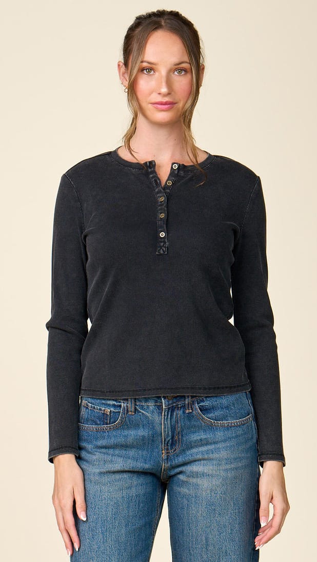A woman modeling a dark charcoal gray long-sleeve henley shirt with blue jeans, standing against a solid light beige background.