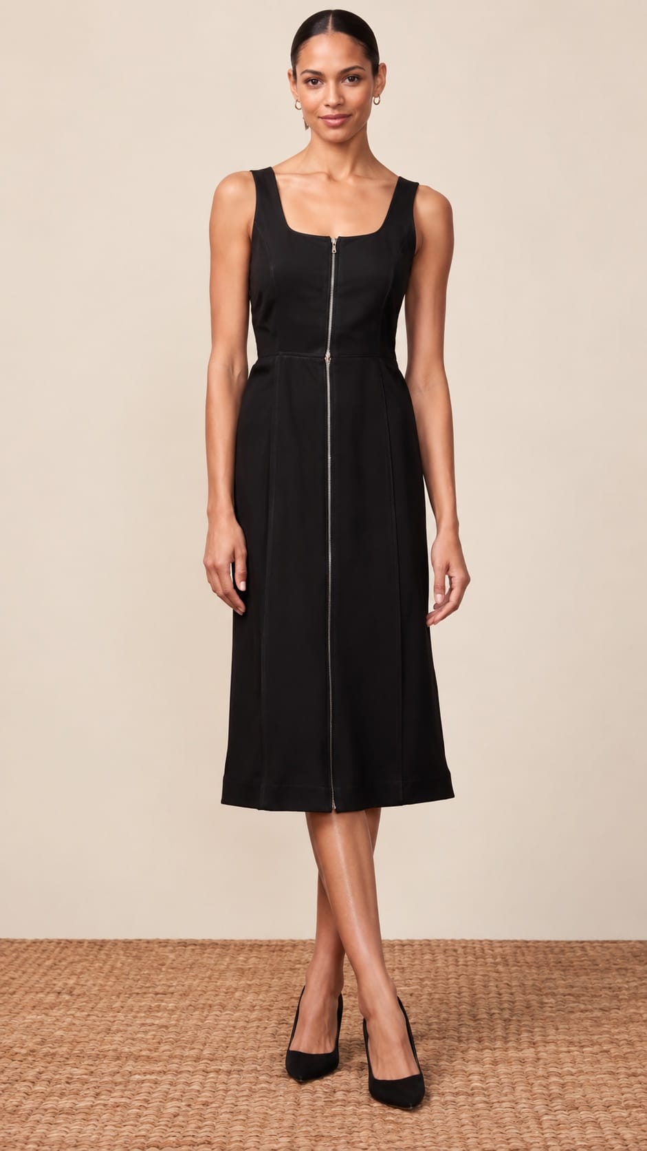 A woman modeling a sleeveless black midi dress with a square neckline, a central front zipper, and a flared skirt, standing against a plain beige studio backdrop.