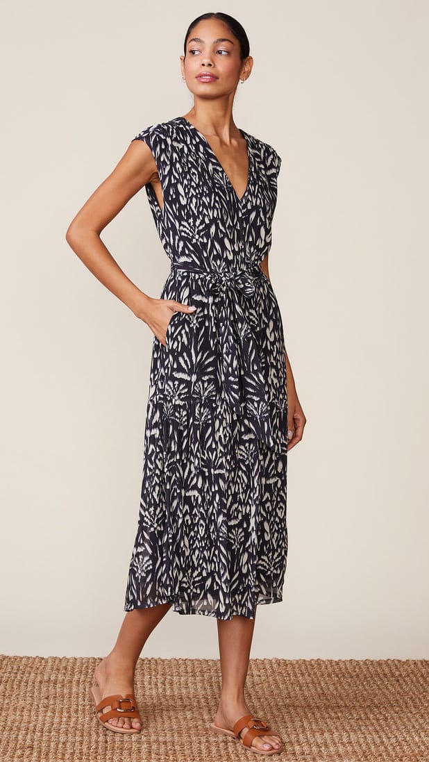 A woman modeling a sleeveless midi wrap dress with a black and white botanical leaf pattern, standing on a woven jute rug against a plain beige background.