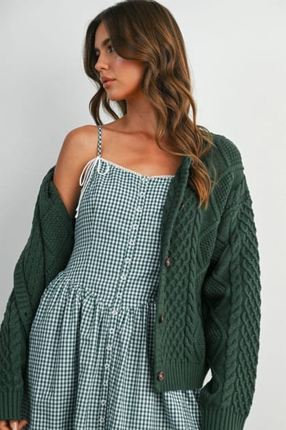 A woman wearing a green and white gingham dress with a dark green cable-knit cardigan, photographed against a plain light gray studio background.