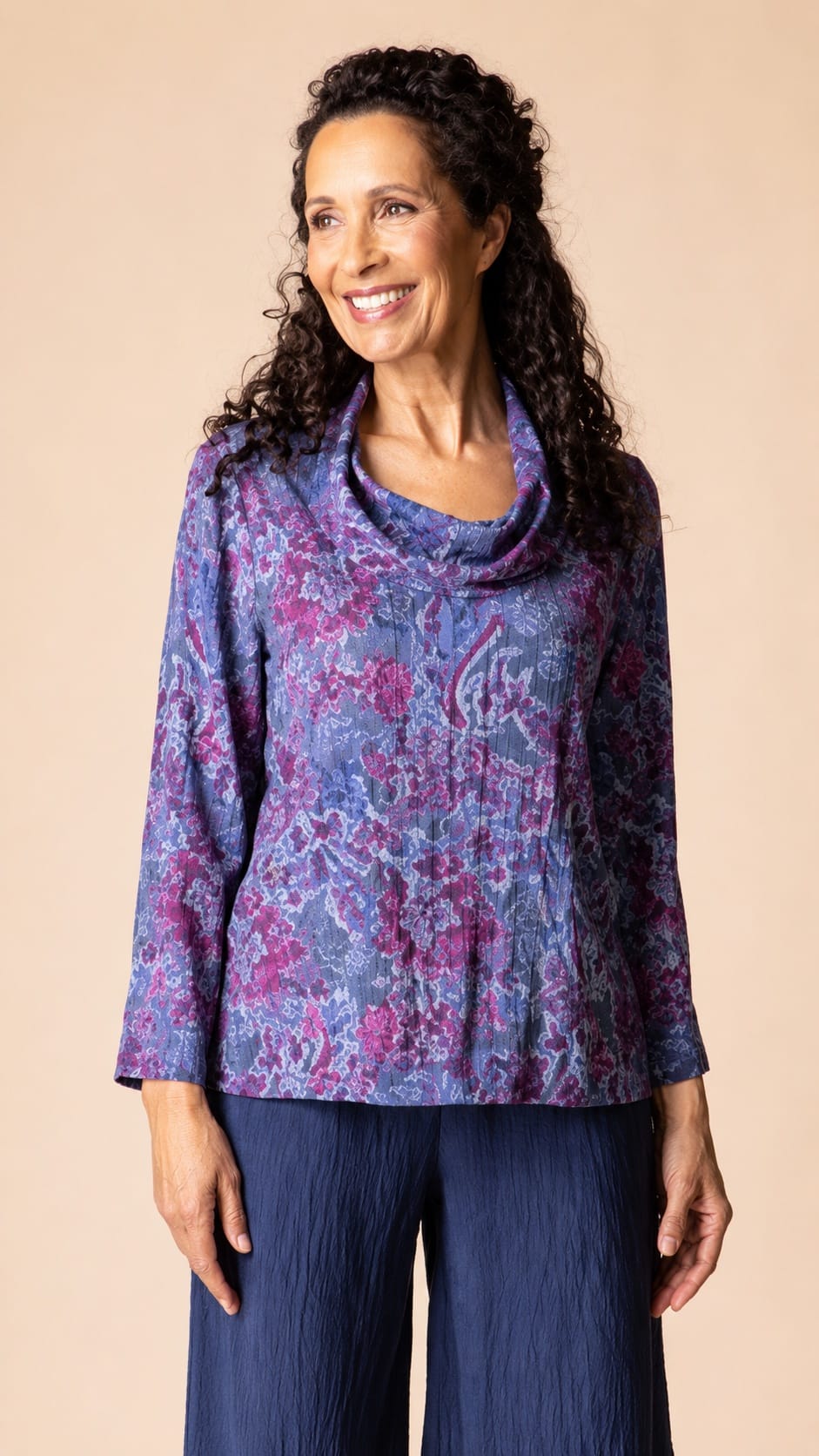 A woman with long curly dark hair wearing a purple and blue patterned cowl-neck top and dark blue pants, standing against a solid beige background.