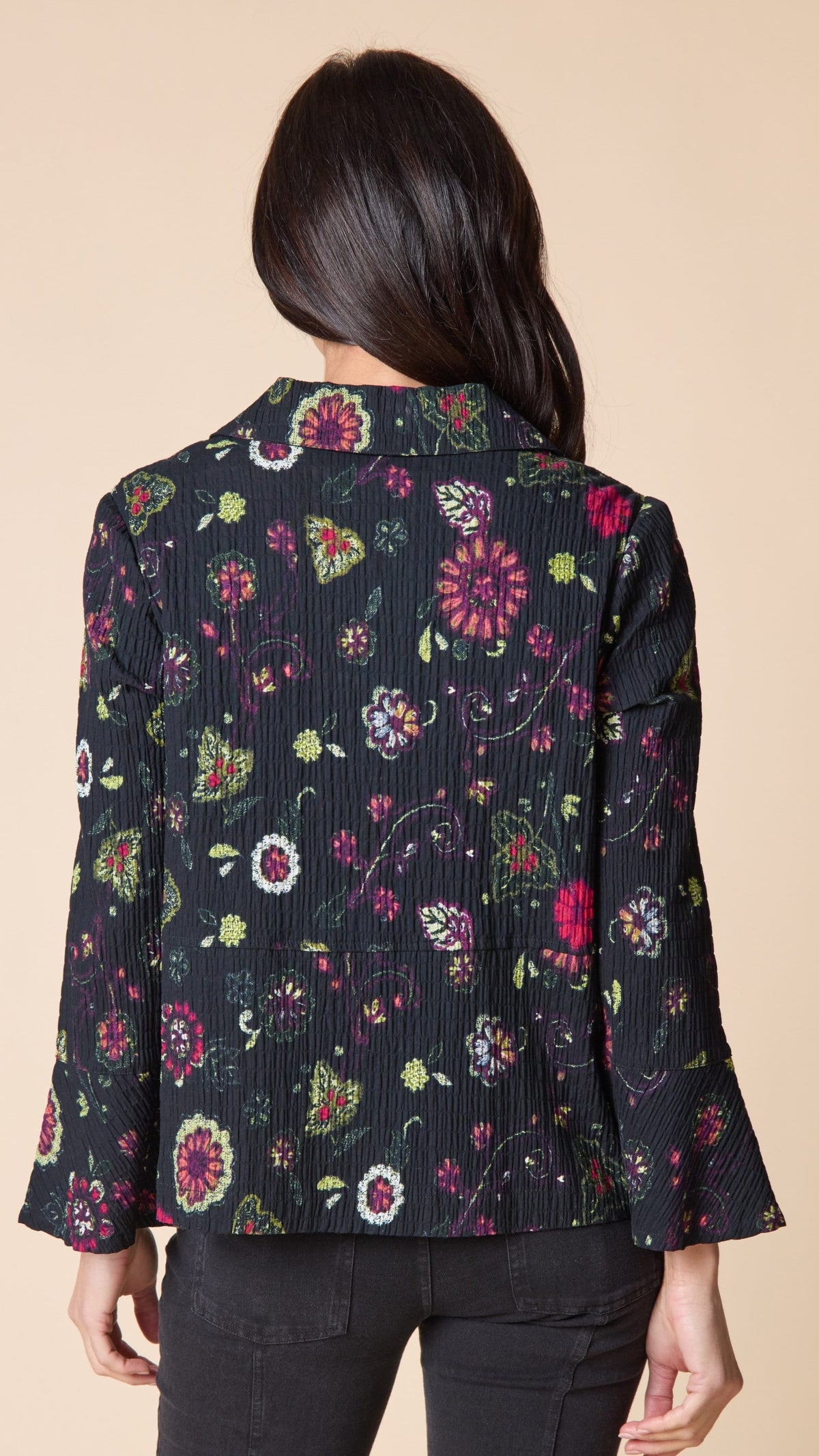 A woman shown from the back wearing a dark pleated floral jacket with pink, red, and green flowers, paired with black pants, against a solid beige background.