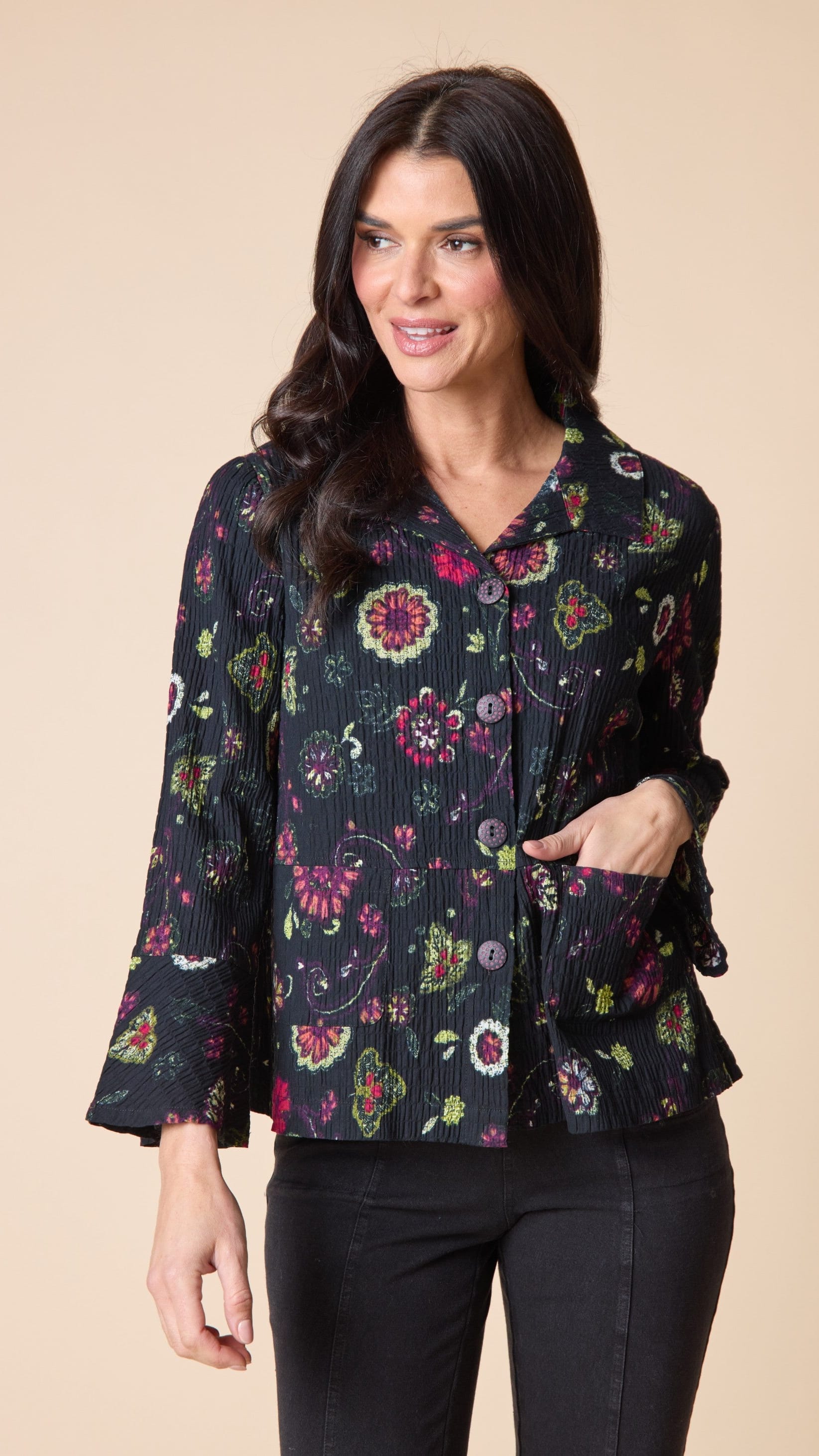 A woman modeling a black long-sleeve blouse with a vibrant multicolor floral print, pleated texture, and side pockets, paired with black pants against a solid beige background.
