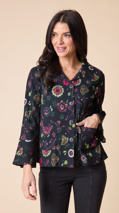 A woman modeling a black long-sleeve blouse with a vibrant multicolor floral print, pleated texture, and side pockets, paired with black pants against a solid beige background.