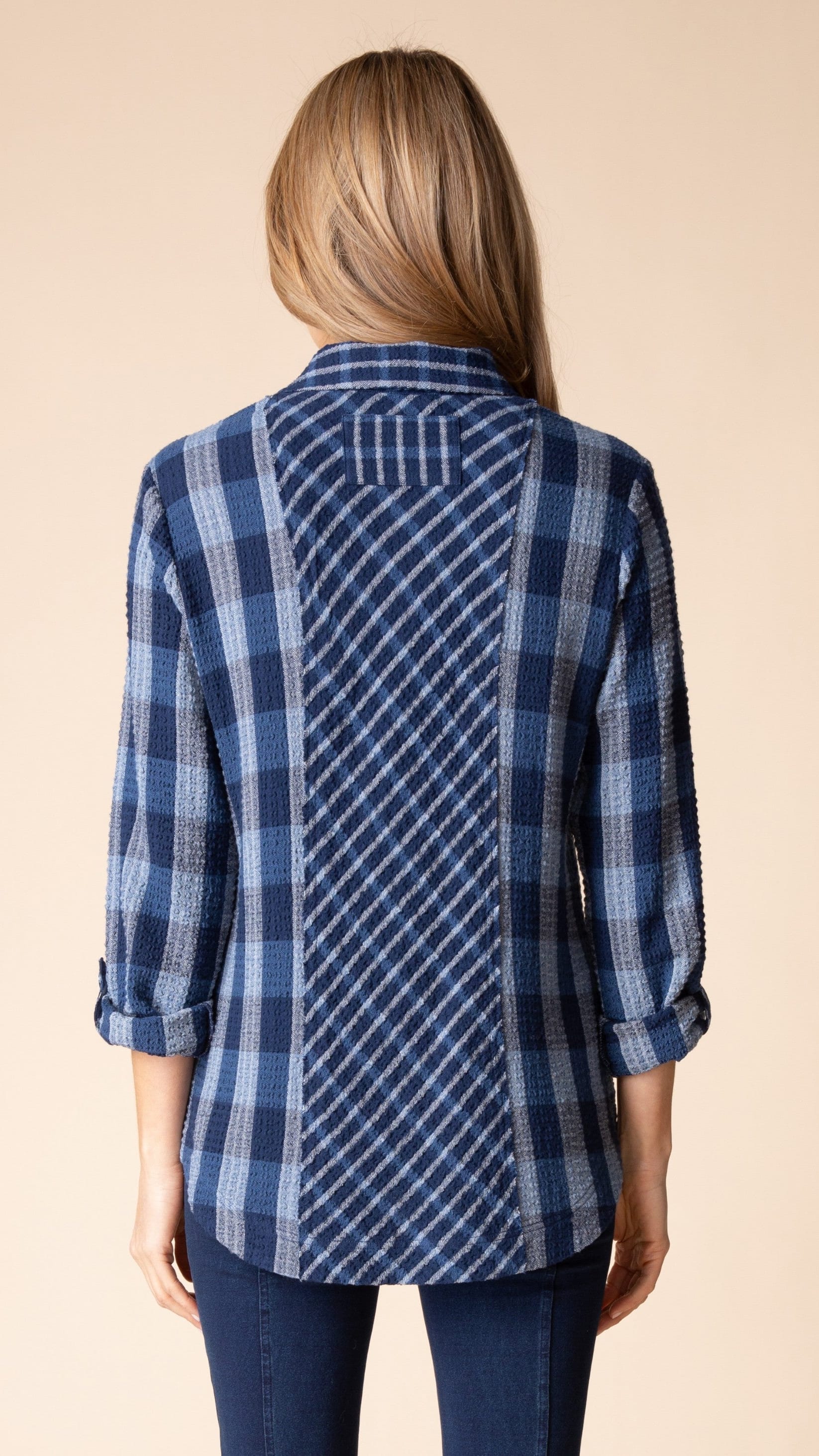 A woman viewed from the back wearing a blue and navy plaid button-up shirt with a unique textured panel design on the back, paired with dark blue jeans, against a solid beige background.
