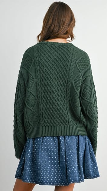 A woman shown from the back wearing a dark green cable-knit sweater and a blue polka-dot skirt against a plain light background.