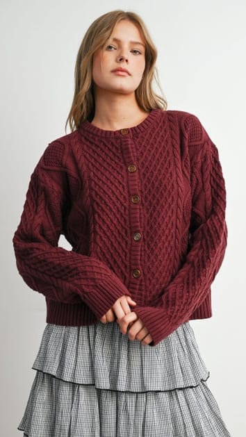 A woman wearing a chunky cable-knit cardigan in deep burgundy with brown buttons, paired with a gray and white checkered tiered skirt, against a plain light gray background.