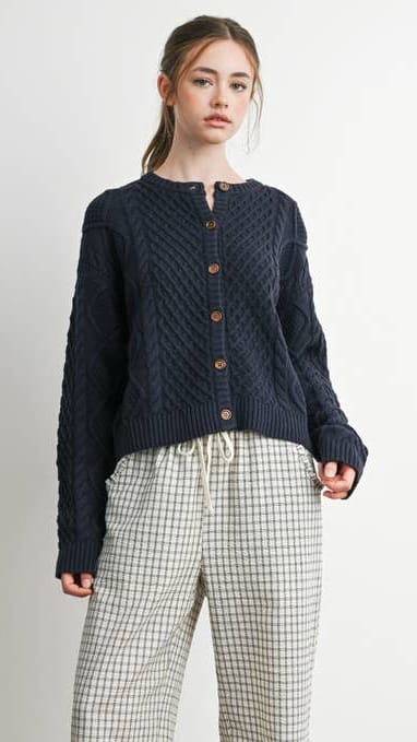 A woman modeling a dark navy cable-knit cropped cardigan with brown buttons, paired with beige and black checkered wide-leg pants, against a plain off-white background.
