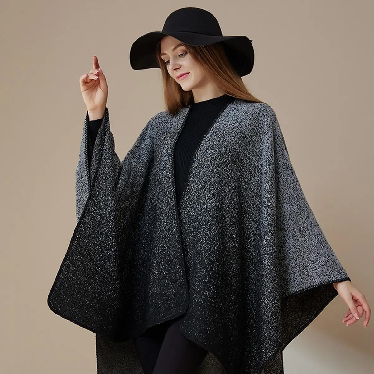 A woman wearing a black wide-brim hat, a black top, and a black-and-gray speckled poncho-style cape against a plain beige studio background.