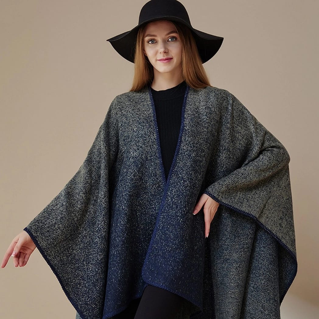 A woman wearing a dark gray and navy blue heathered poncho-style wrap over a black top, with a wide-brim black hat, posed against a plain beige studio background.