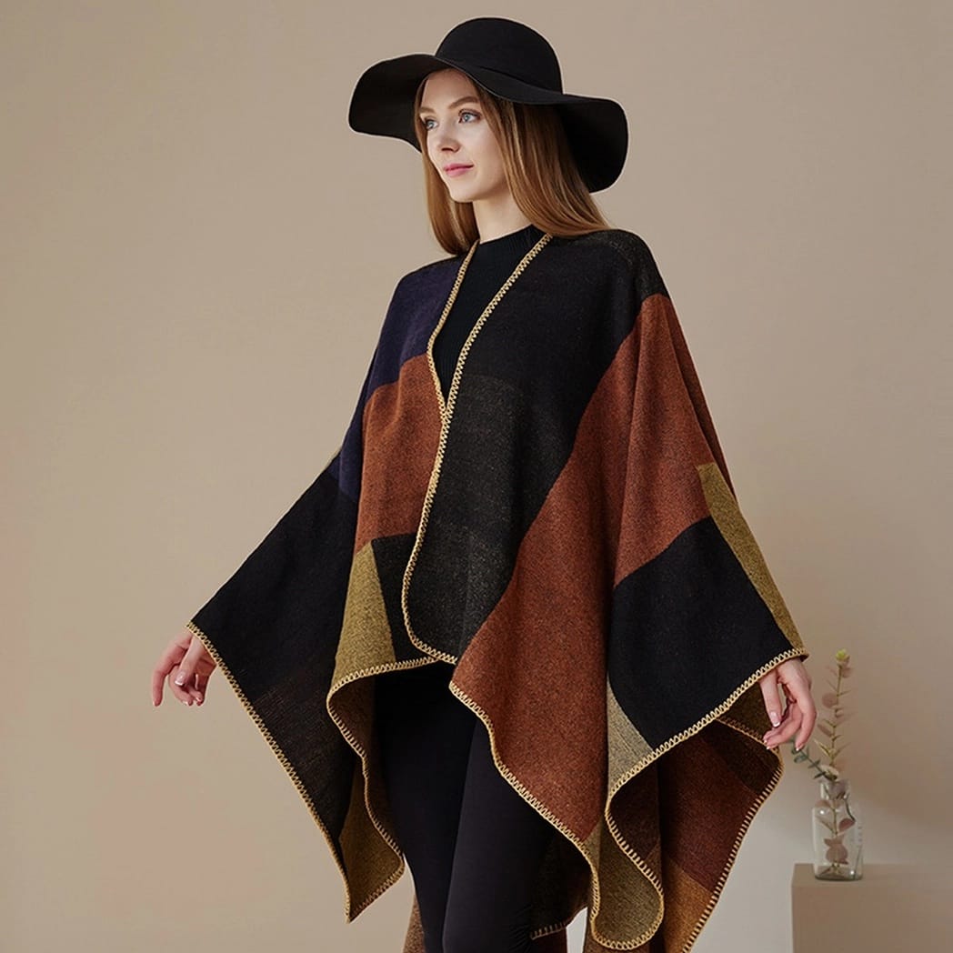 A woman wearing a large black wide-brim hat and a multi-colored patchwork poncho with brown, black, and gold sections, standing against a neutral beige background.