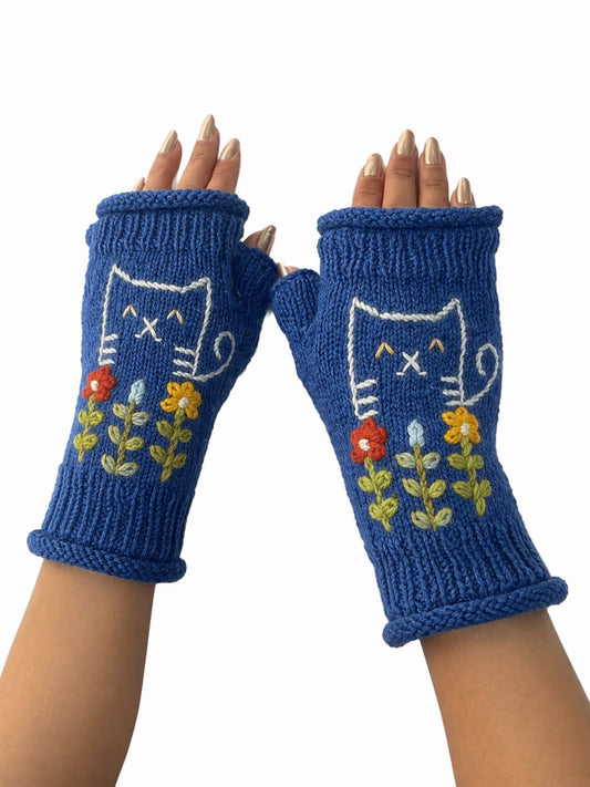 A pair of blue fingerless knit gloves with a white cat face and colorful embroidered flowers, shown on hands against a plain white background.