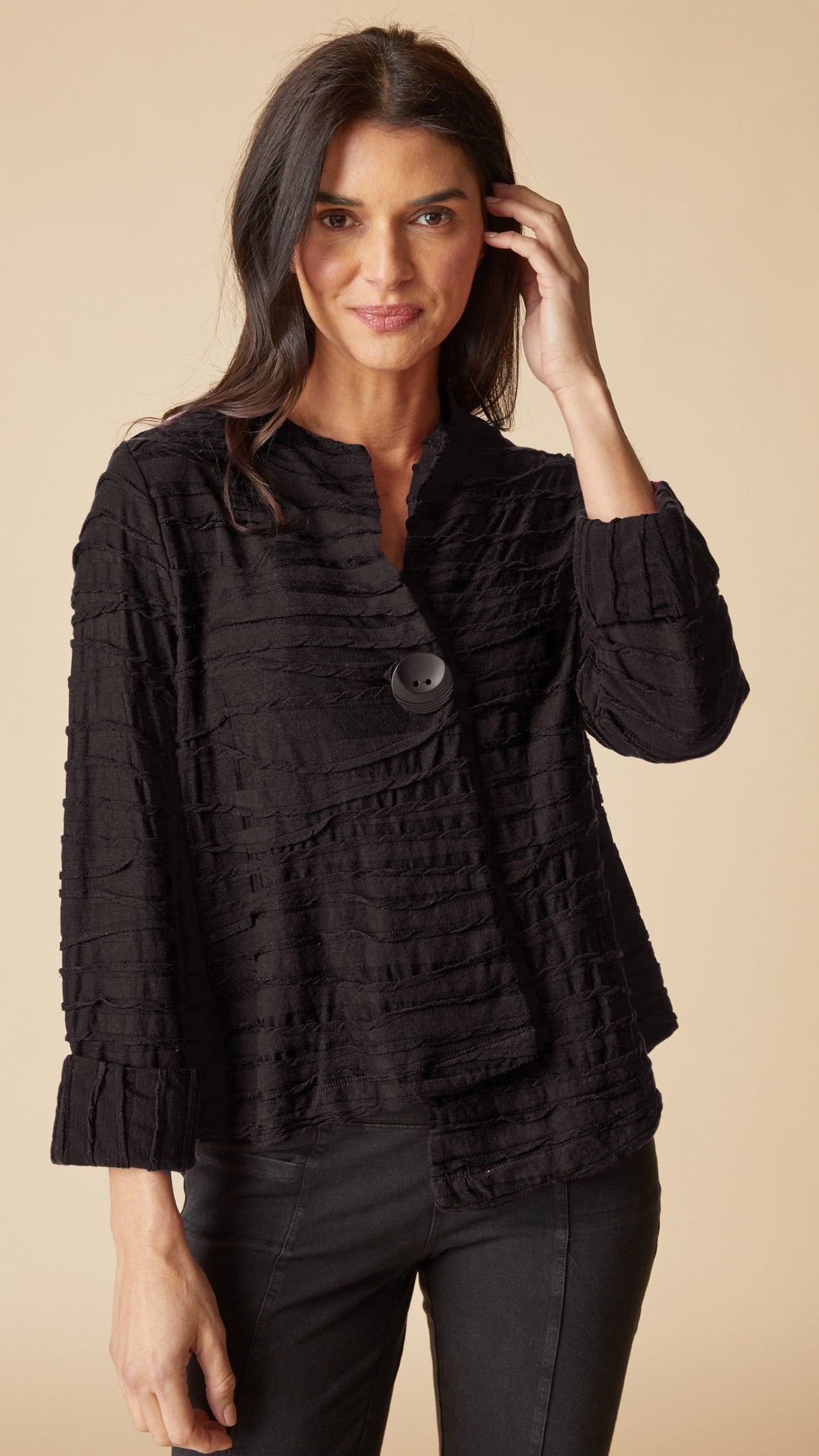 A woman wearing a black textured, ruffled long-sleeve blouse with a buttoned collar, paired with black pants, set against a solid beige background.
